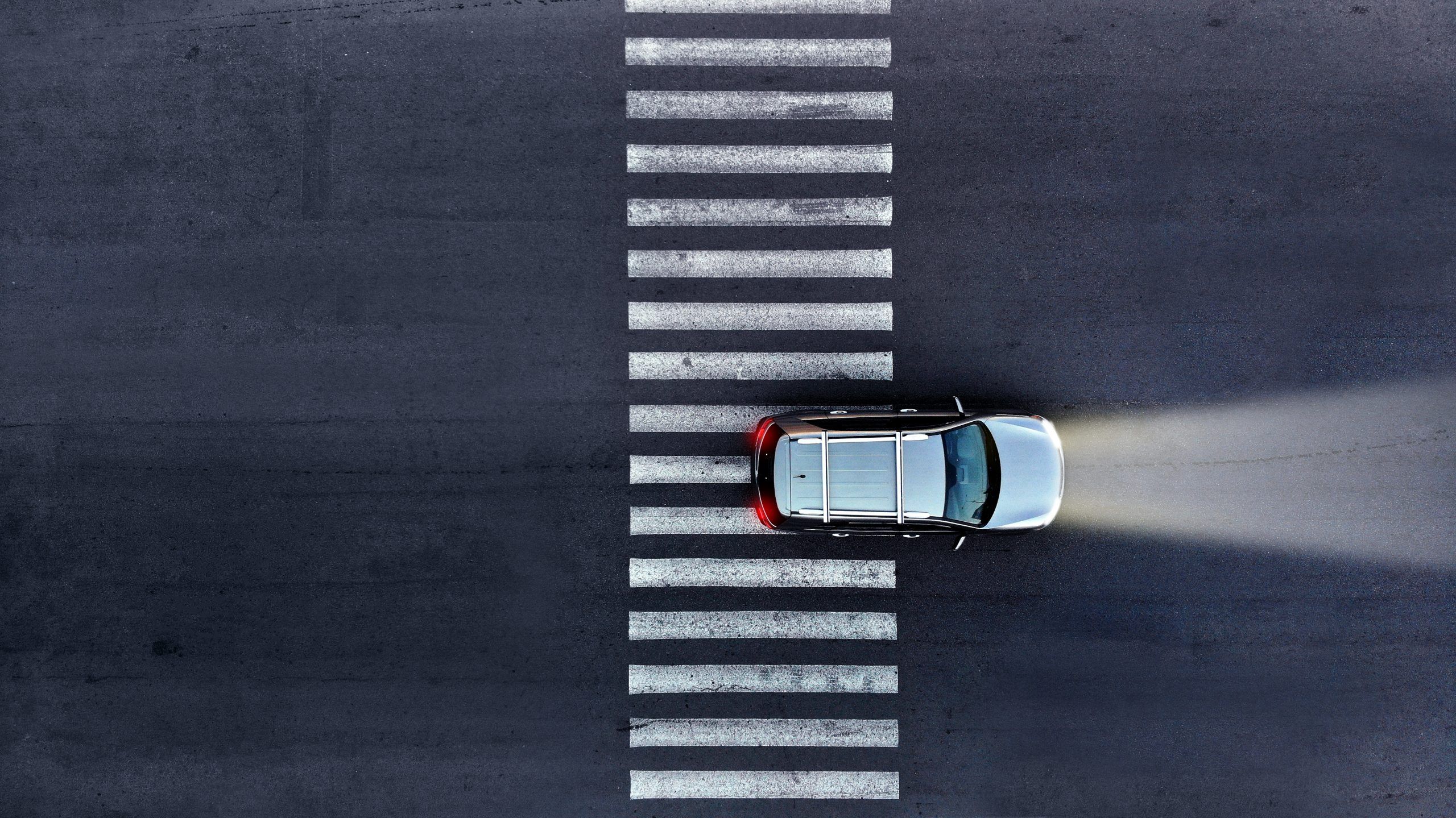 An aerial view of a silver car on a zebra crossing, its high beams are on